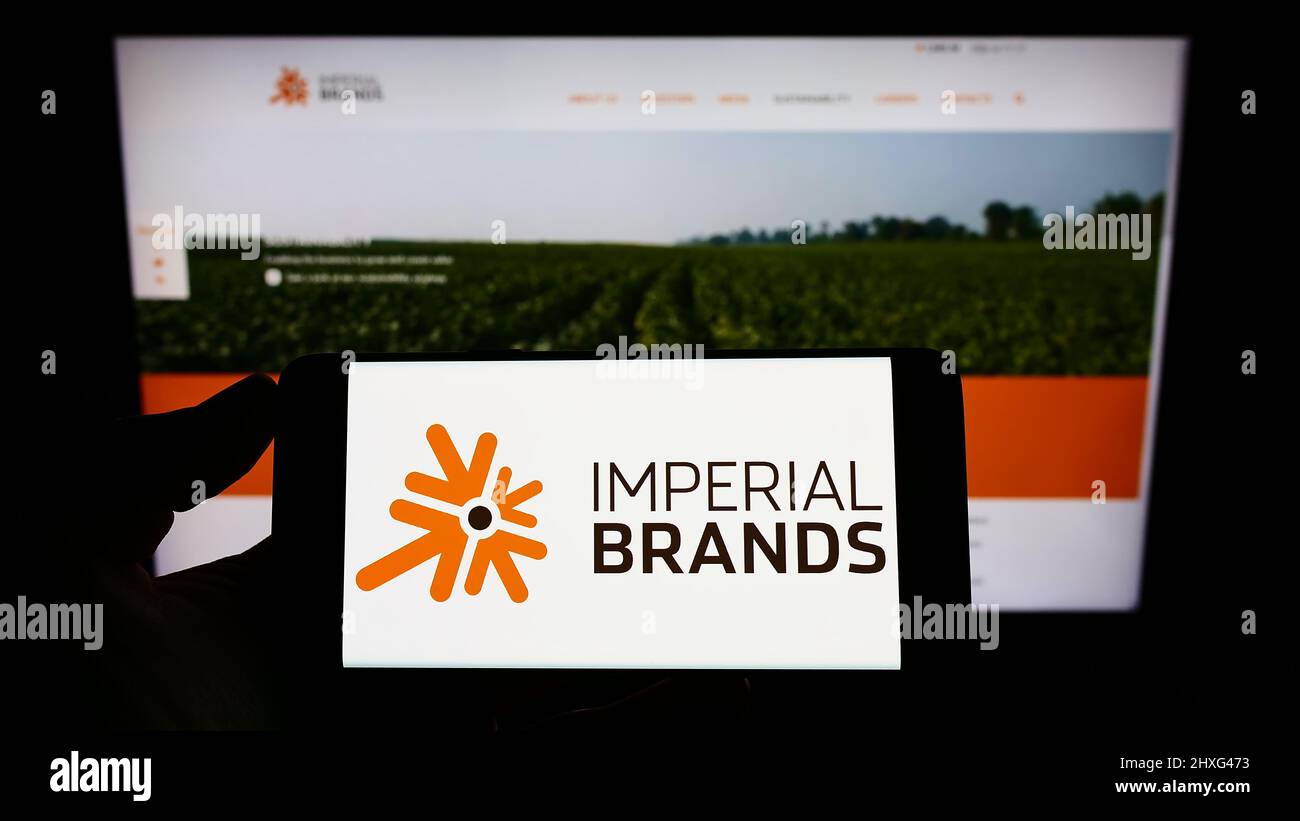Persona che tiene uno smartphone con il logo della società britannica di tabacco Imperial Brands plc sullo schermo di fronte al sito web. Mettere a fuoco sul display del telefono. Foto Stock