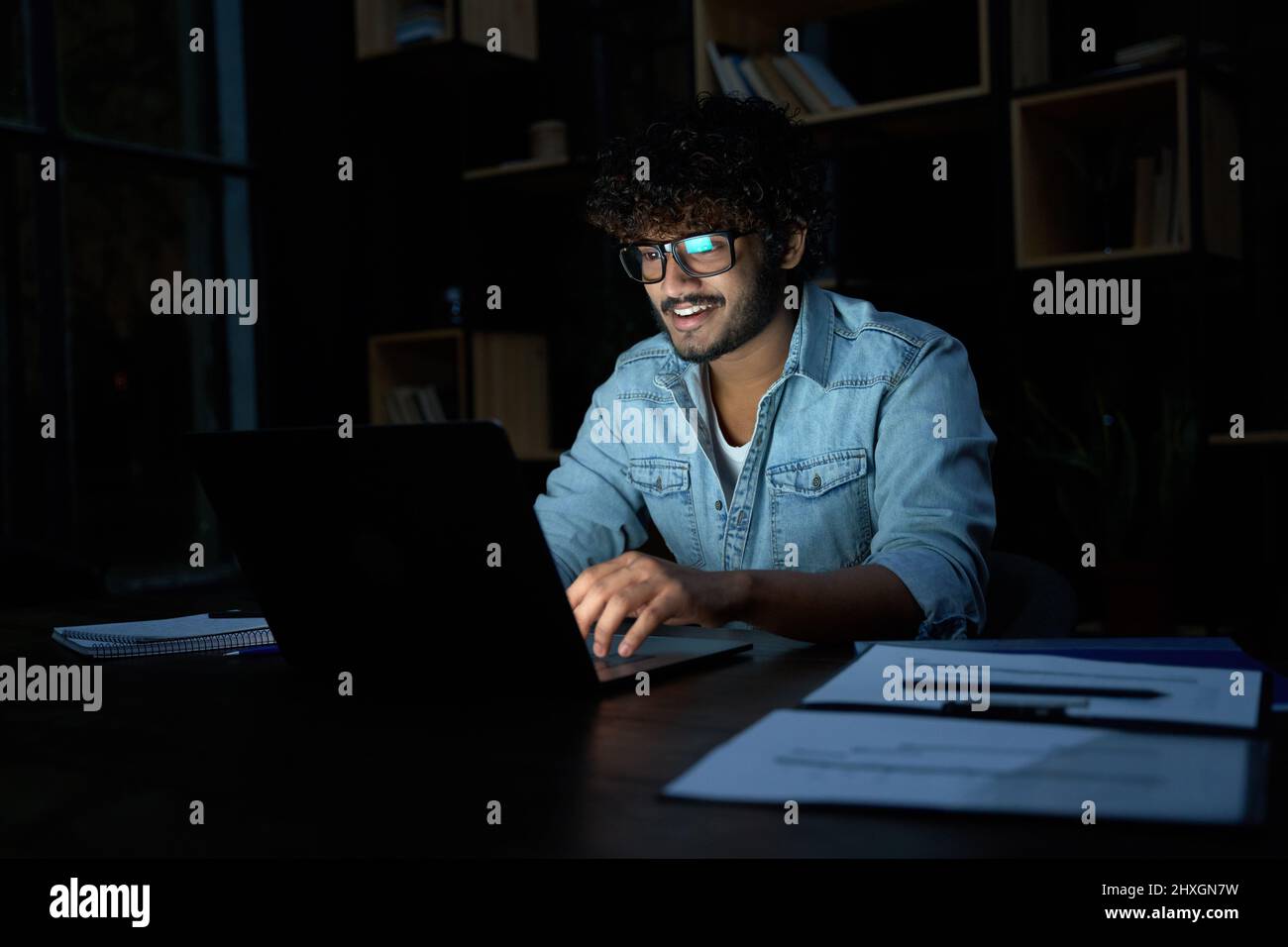 Sorridente giovane uomo d'affari indiano che lavora in linea a tarda notte usando il laptop. Foto Stock