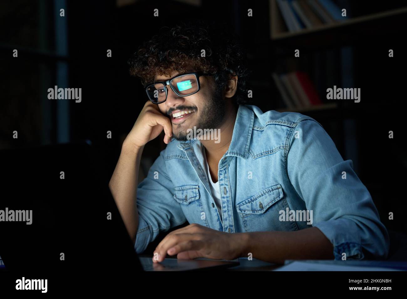 Felice uomo indiano giovane che lavora in linea a tarda notte usando il computer portatile. Foto Stock