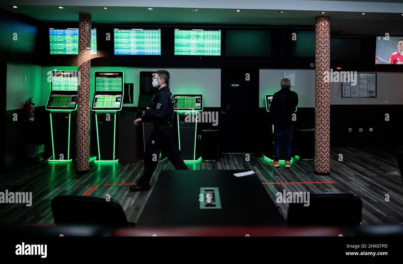 Duisburg, Germania. 12th Mar 2022. Un ufficiale di polizia cammina attraverso un negozio di scommesse mentre un uomo sta ancora operando una delle macchine. La polizia raid un negozio di scommesse nel nord di Duisburg. Ministro degli interni della Renania settentrionale-Vestfalia, Herbert Reul (CDU) è sul posto per ottenere un quadro della situazione. Credit: Fabian Strauch/dpa/Alamy Live News Foto Stock