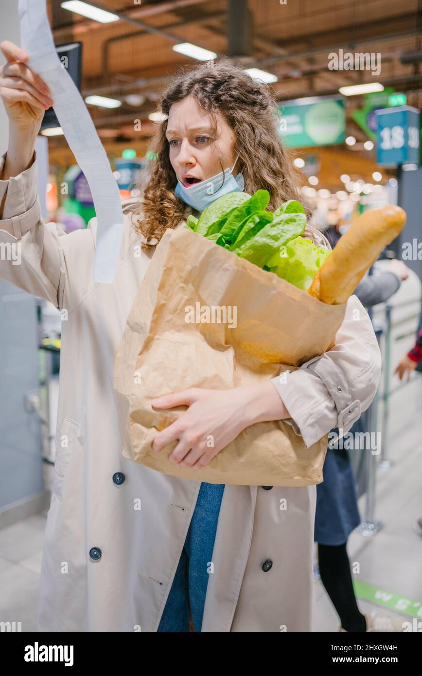 La giovane donna è sorpresa spiacevolmente mentre osserva in un controllo della carta in un supermercato della drogheria e tiene una borsa della spesa. Aumento dei prezzi dei prodotti alimentari. Foto Stock