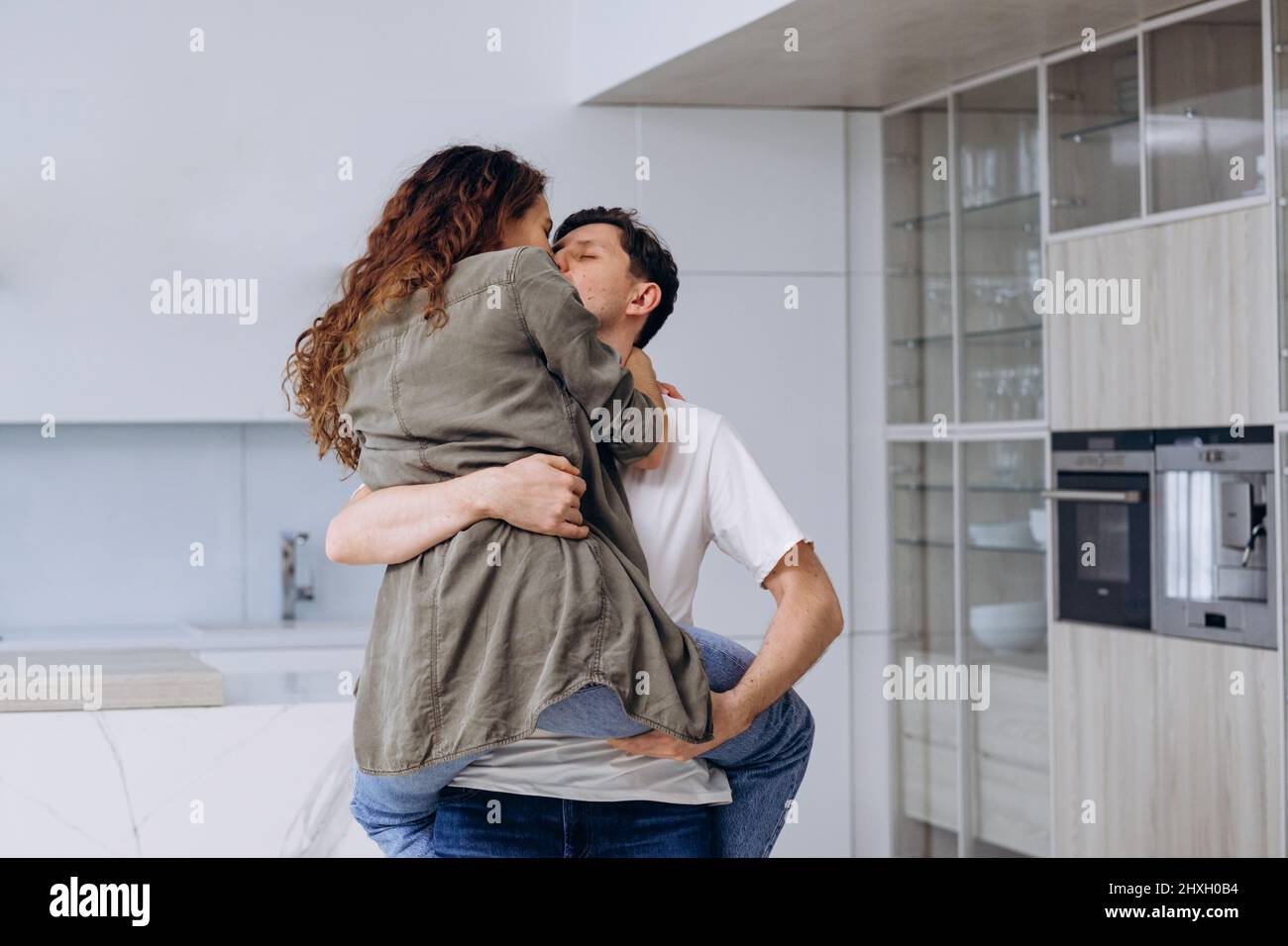 Giovane uomo brunetto cattura la tenuta in braccia e gira ricci moglie in camicia e jeans abbracciando strettamente e ridendo fortunatamente in cucina primo piano Foto Stock