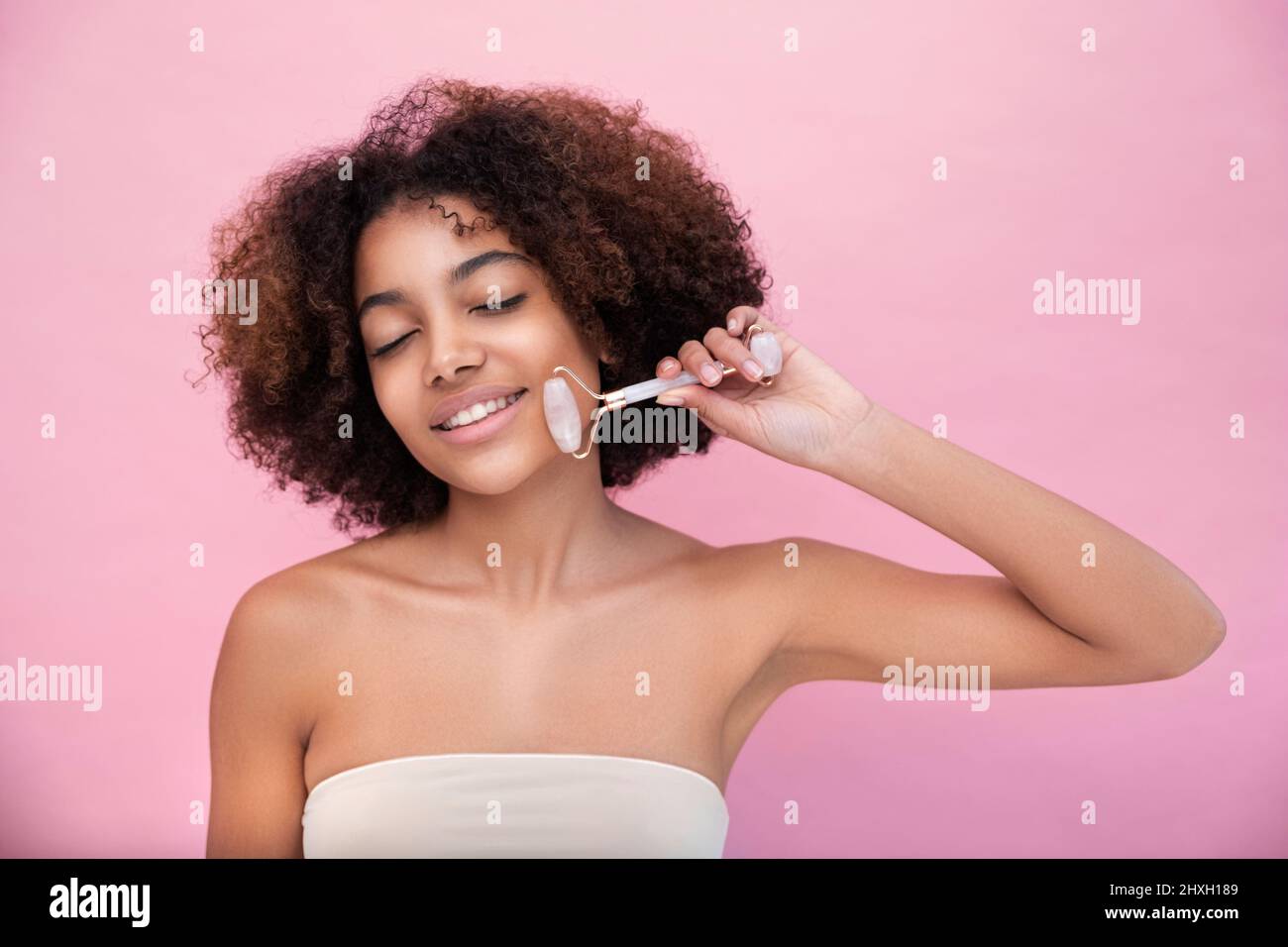 Ritratto di una giovane donna ben curata con pelle scura e capelli lussureggianti, facendo massaggio facciale con l'aiuto di un rullo con occhi chiusi su sfondo rosa primo piano, cura e cura della pelle Foto Stock