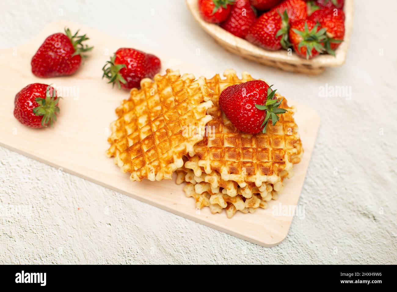 Waffle serviti con fragole su un tagliere bianco su un tavolo grigio e un cestino con fragole mature Foto Stock