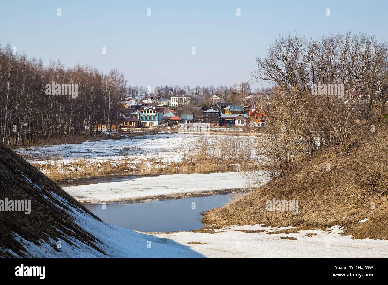 Un fiume ghiacciato che si estende in lontananza, case di legno in lontananza. Primavera, neve si scioglie, pozzanghere e erba secca tutto intorno. Giorno, tempo nuvoloso, luce calda e soffusa. Foto Stock