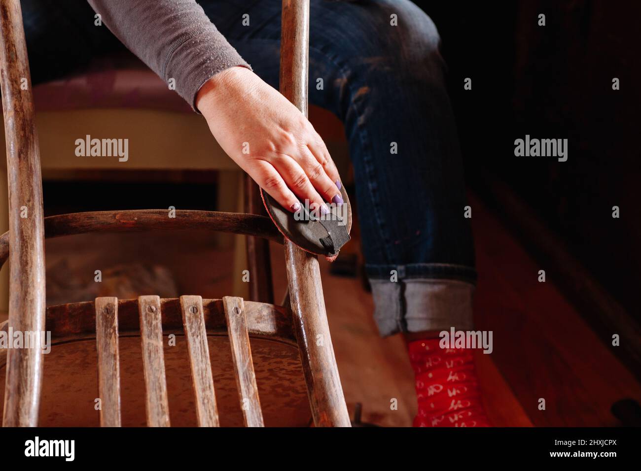 Primo piano di seduta donna levigatura superficie di vecchio sgabello che ha bisogno di ristrutturazione con pezzo di carta vetrata in casa officina. Pratiche sostenibili. Riutilizzo del vecchio Foto Stock