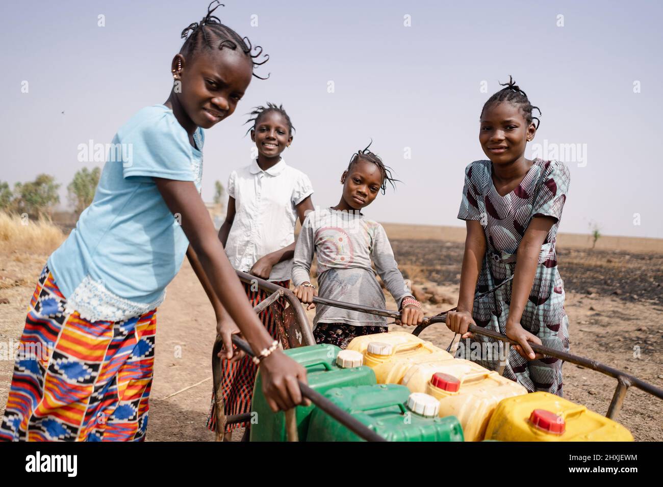 Quattro ragazze africane graziose che spingono insieme un carrello pieno di lattine d'acqua pesanti; simbolico per l'Unione è la forza; concetto di lavoro infantile Foto Stock