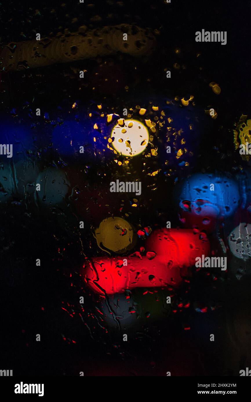 Scatto astratto di gocce di pioggia su una finestra di vetro, con luci sullo sfondo sfocate per creare un effetto bokeh brillante, aggiungendo profondità. Foto Stock