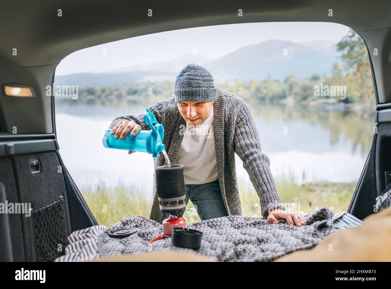 Un uomo sorridente vestito con abiti caldi a maglia versando un'acqua dolce in stufa a gas nel comodo bagagliaio dell'auto con una splendida vista sul lago di montagna. Caldo Foto Stock