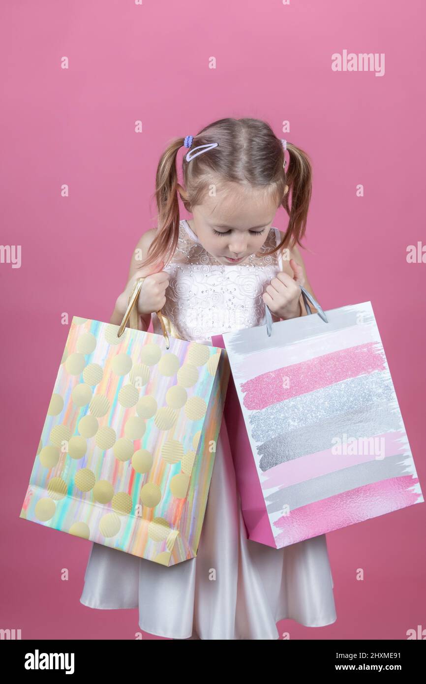 Carino ragazza caucasica piccola in un abito da festa su sfondo rosa felice godendo shopping tenendo borse colorate Foto Stock