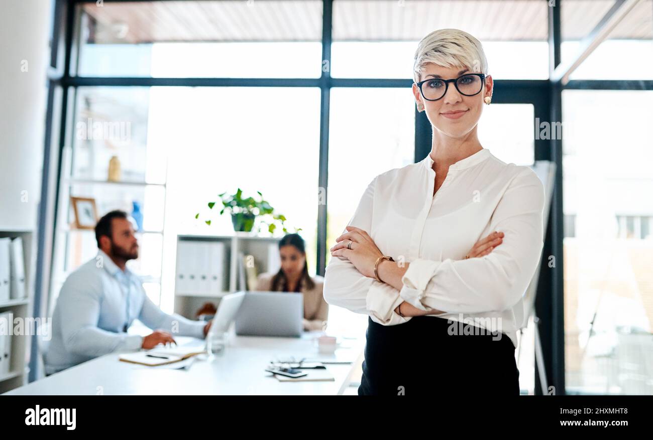 Posso essere una mamma e un go-getter tutto in uno. Ritratto di una donna d'affari incinta in piedi in un ufficio con i suoi colleghi in background. Foto Stock