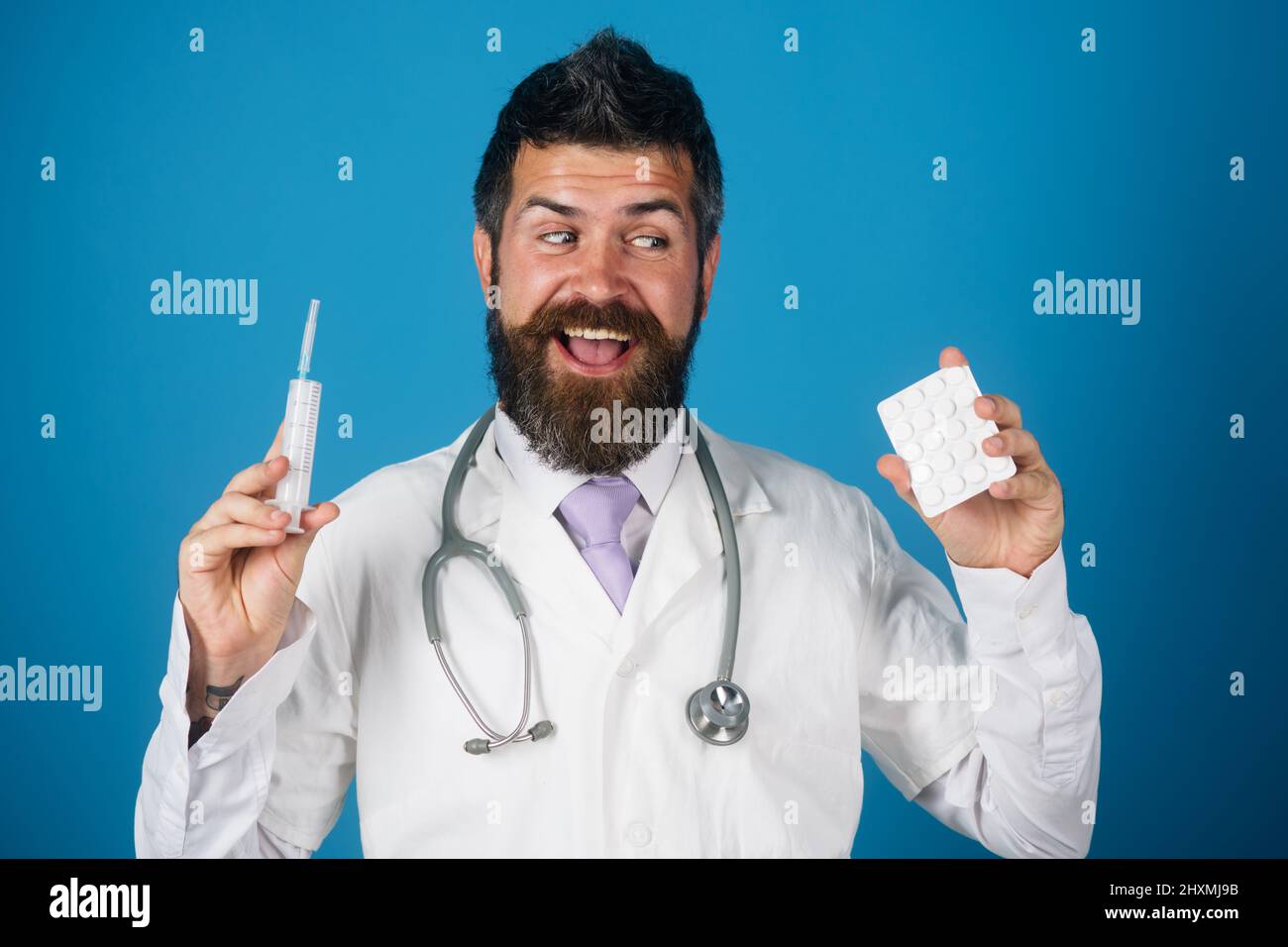 Dottore in bearded con pillole e siringa in mani. Medico sorridente con stetoscopio e abito medico. Foto Stock