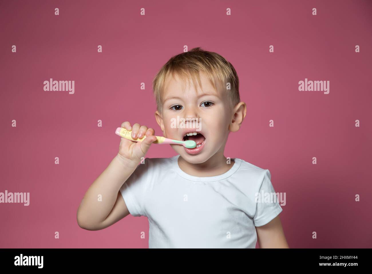 Sorridente ragazzino caucasico che pulisce i denti con lo spazzolino manuale per bambini su sfondo rosa. Foto Stock