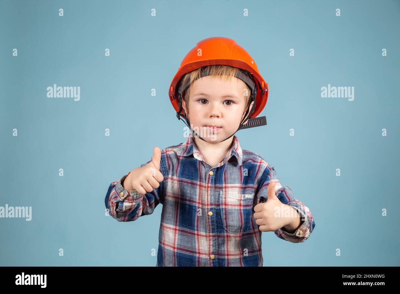 Divertente piccolo ragazzo ingegnere o architetto in un casco arancione protettivo su sfondo blu mostrando il pollice in su. Concetto di costruzione e riparazione. Foto Stock