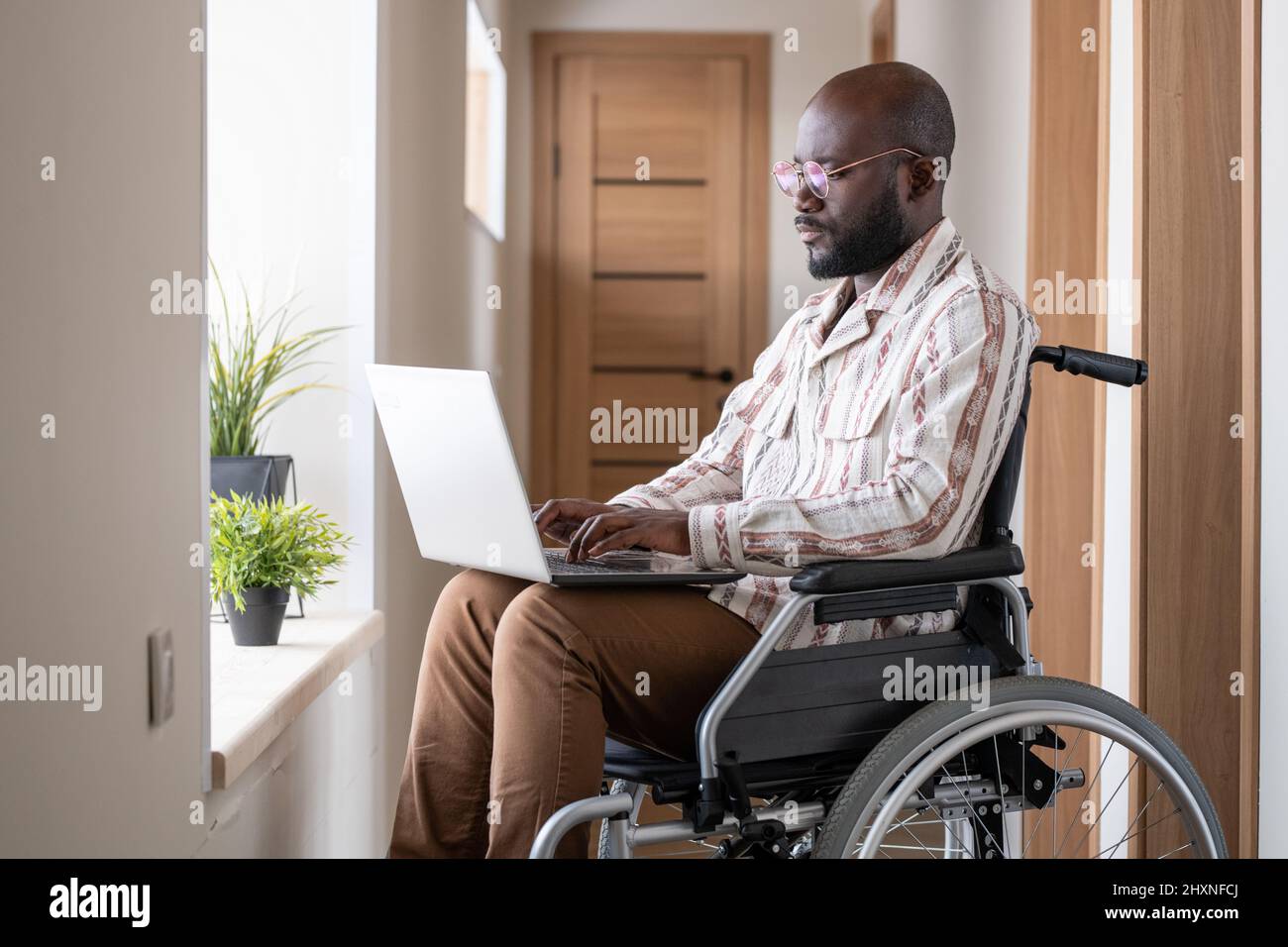 Giovane uomo nero serio con disabilità che si concentra sulla rete mentre si siede in sedia a rotelle nel corridoio dell'appartamento Foto Stock