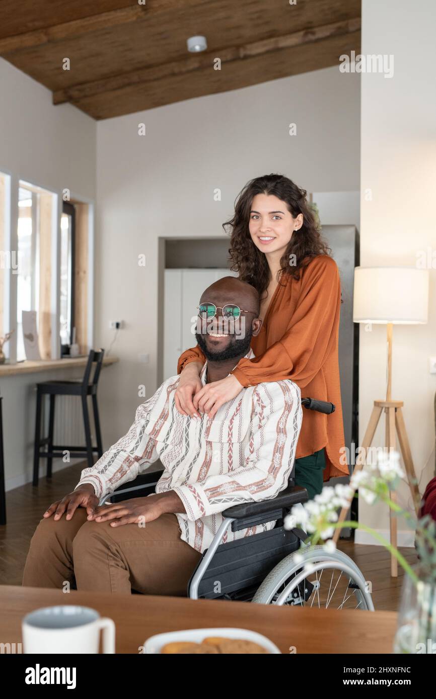Felice giovane coppia interrazziale in casualwear guardando la macchina fotografica con sorrisi mentre si passa il tempo insieme a casa Foto Stock