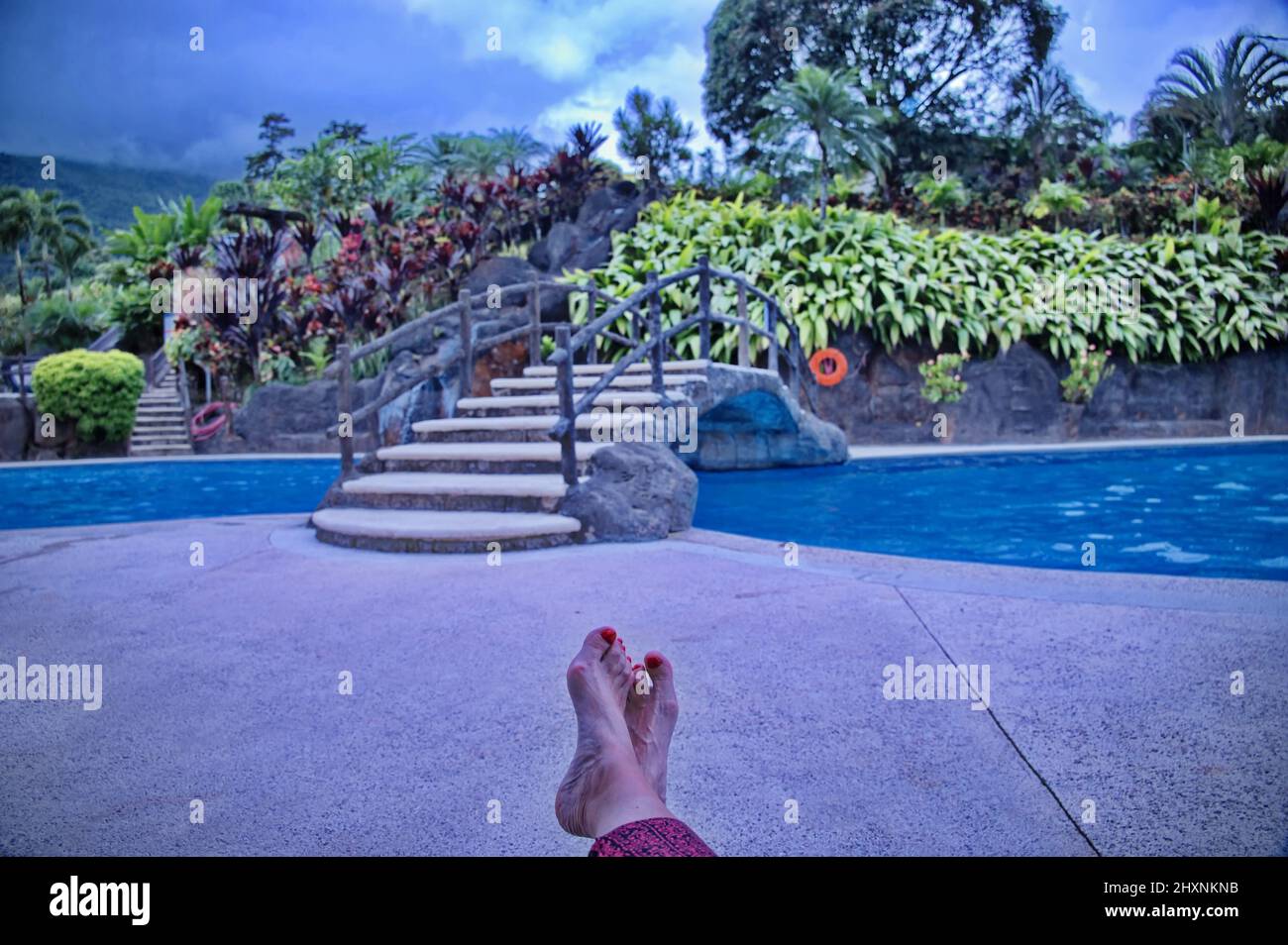 Gambe femminili senior con unghie rosse di fronte alla piscina con piante tropicali Foto Stock