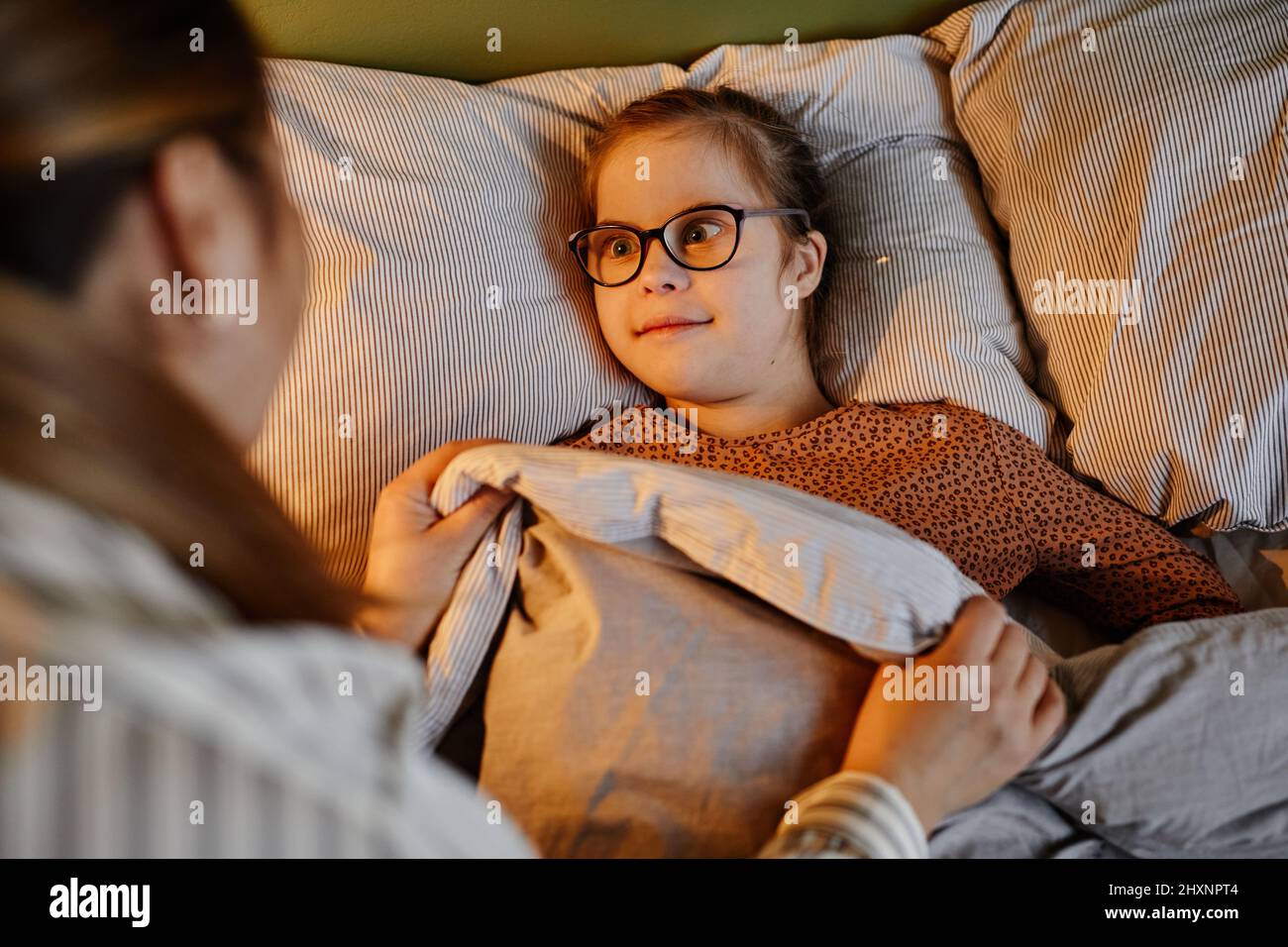 Ritratto ad angolo alto di cura madre che si stucco in bambino con sindrome di Down a letto in luce lampada accogliente, spazio copia Foto Stock