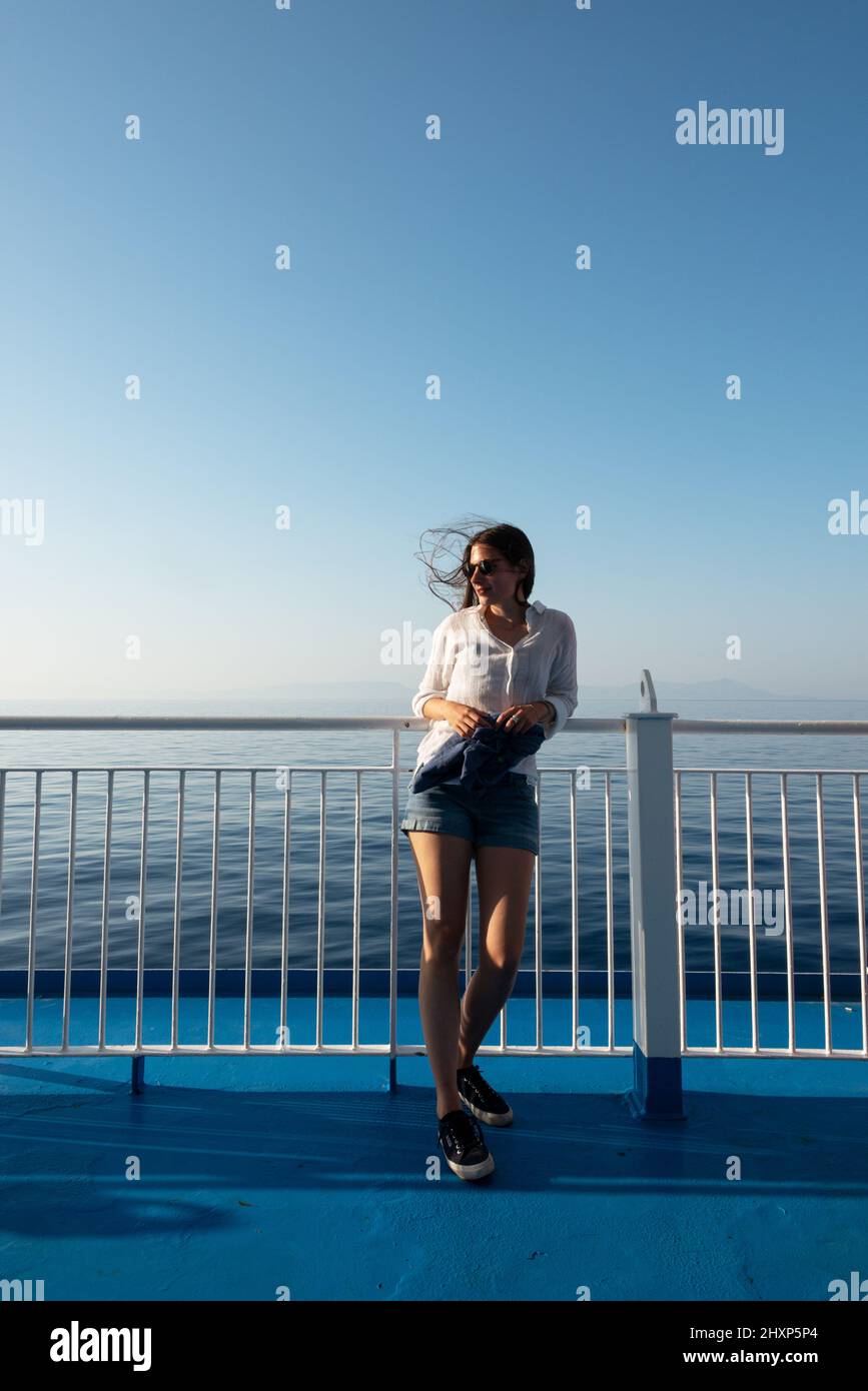 ragazza in pantaloncini con capelli soffiato dal vento appoggiata contro la ringhiera della nave. Foto Stock