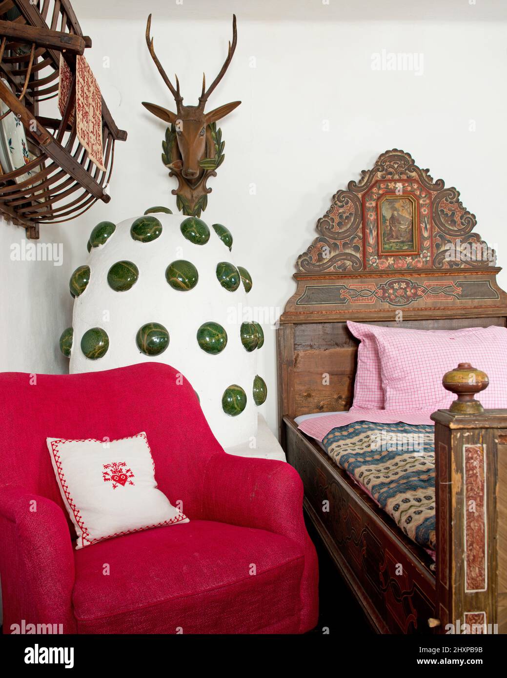 Particolare della camera da letto in stile alpino con letto in legno dipinto, stufa in maiolica in bianco e verde, sedia rossa e corna Foto Stock