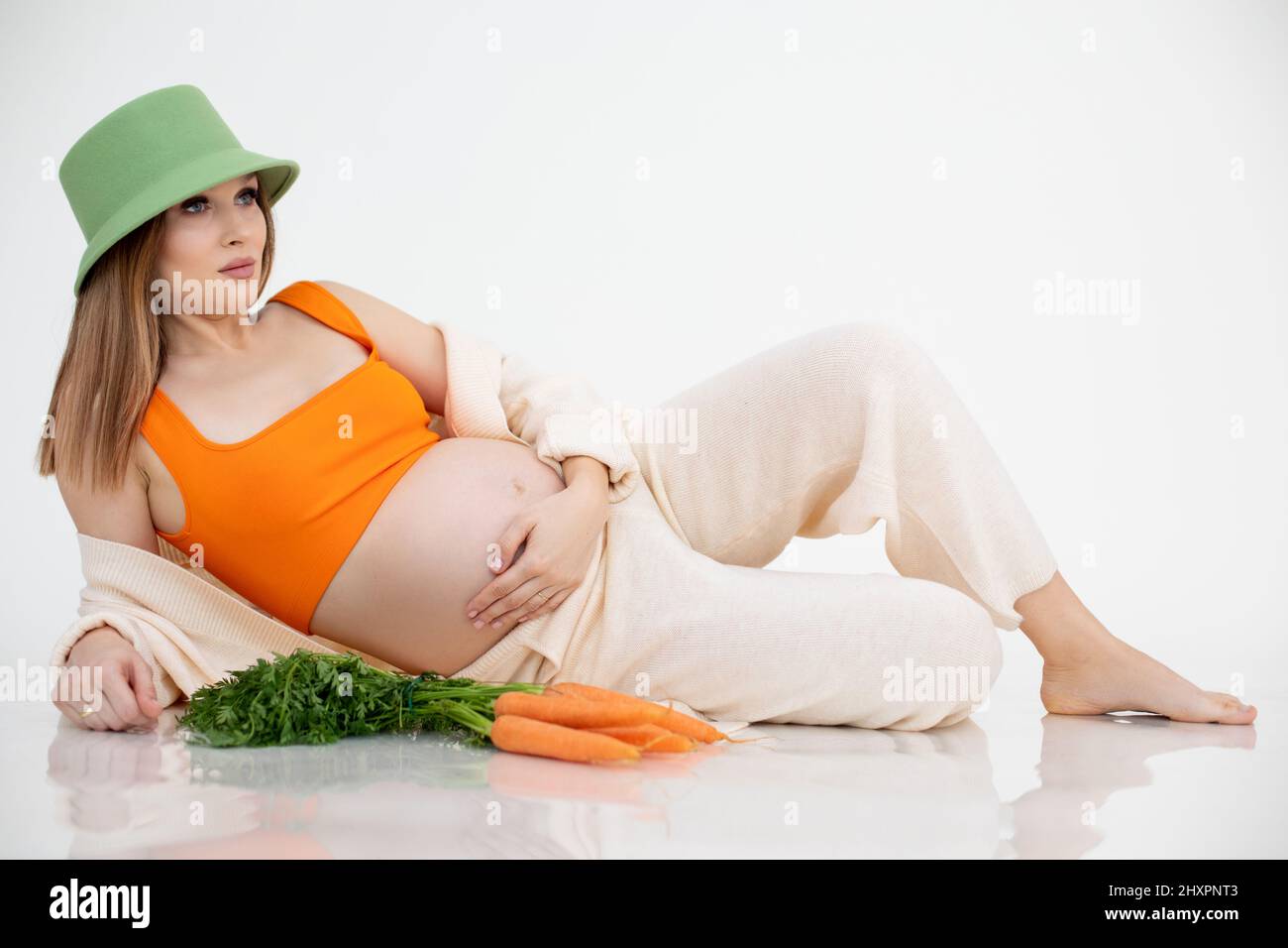 Attraente donna incinta calma a piedi nudi in cappello verde sdraiato, tenere il ventre, mazzo di carote arancio. Stagione del raccolto del giardino. Foto Stock