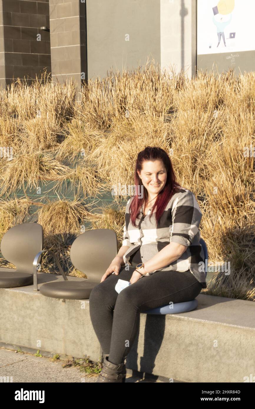 giovane donna seduta su una panca, con una camicia a quadri e capelli rossastri Foto Stock