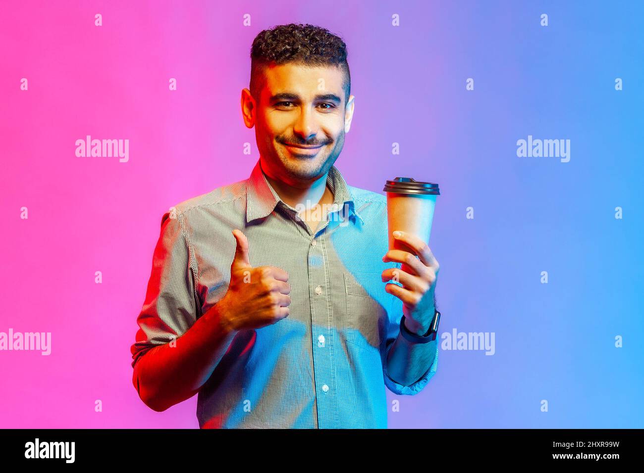 Ritratto di uomo soddisfatto e felice in camicia che tiene il caffè per andare nelle mani e mostrare i pollici in su, guardando la macchina fotografica. Studio interno girato isolato su colorato sfondo di luce al neon. Foto Stock