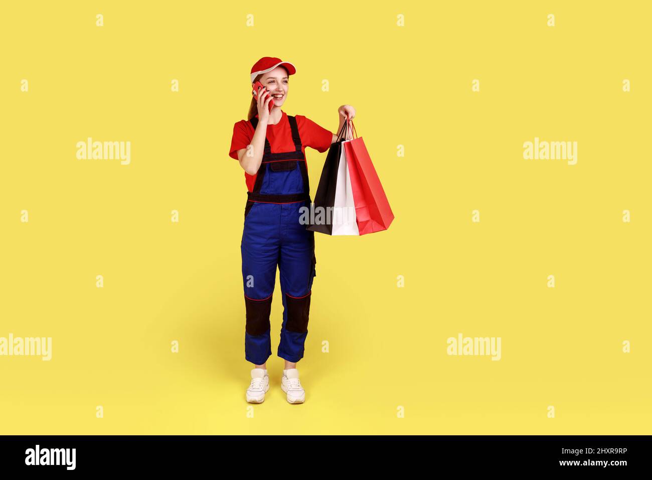 Ritratto a tutta lunghezza di sorridente corriere donna in piedi che tiene i sacchetti di shopping nelle mani, telefono parlante con il cliente con espressione positiva. Studio interno girato isolato su sfondo giallo. Foto Stock