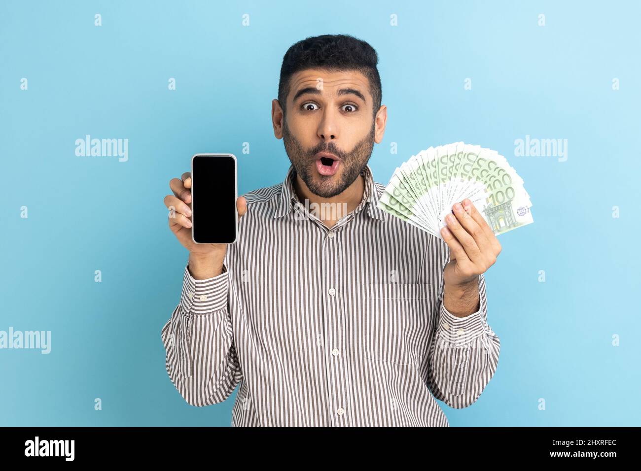 Ritratto di stupito uomo d'affari con grande fan di banconote in euro e telefono cellulare con display vuoto per la pubblicità, indossando maglietta a righe. Studio interno girato isolato su sfondo blu. Foto Stock