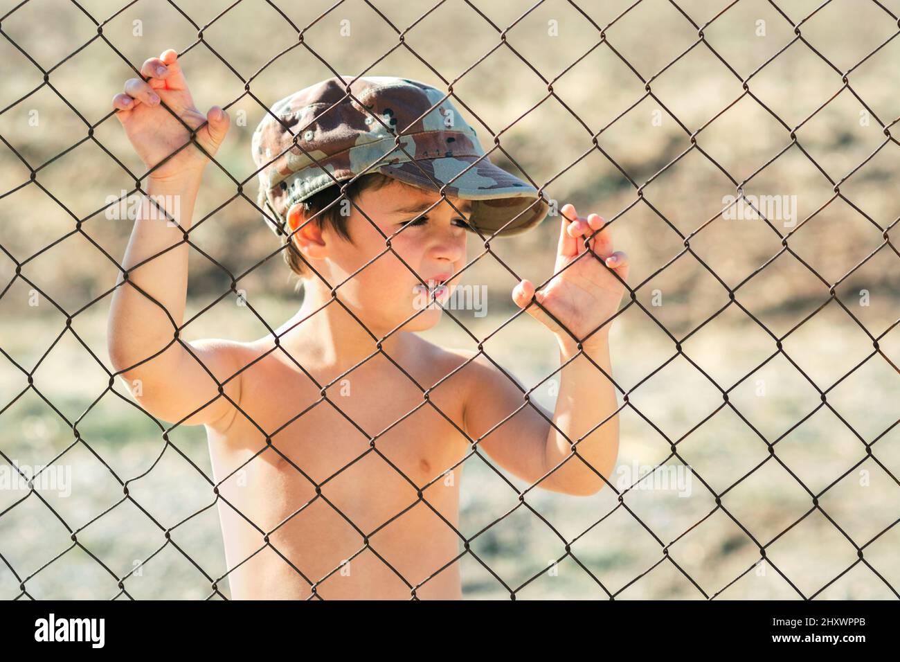 Bambino triste in un tetto militare dietro una recinzione. Arrestare il concetto di guerra Foto Stock