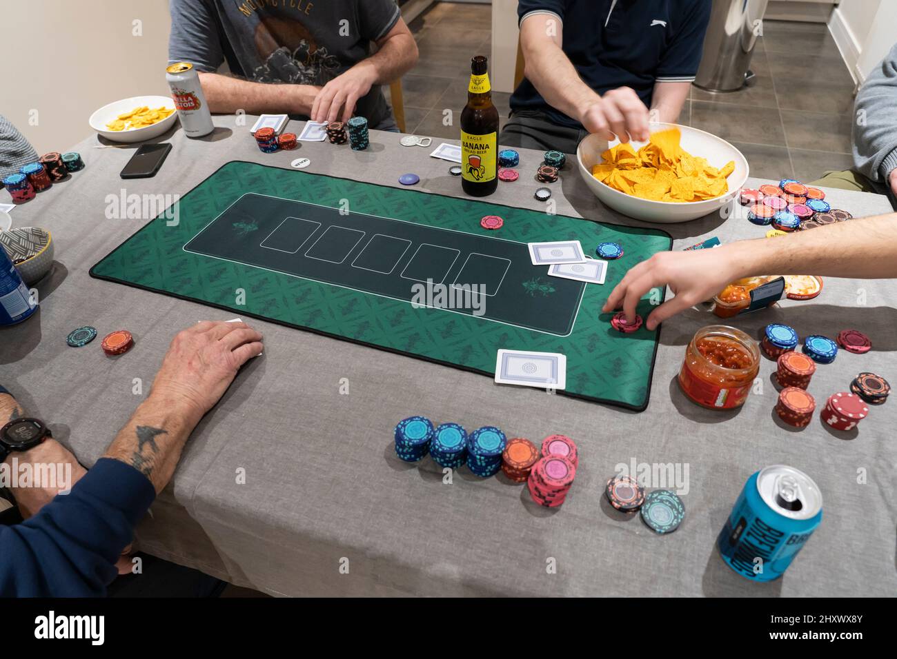 Amici di sesso maschile che giocano a Texas Hold 'em Poker su un tavolo a casa con gioco d'azzardo chip, un mat di poker, patatine e salsa di salsa e birre da bere. Inghilterra Foto Stock
