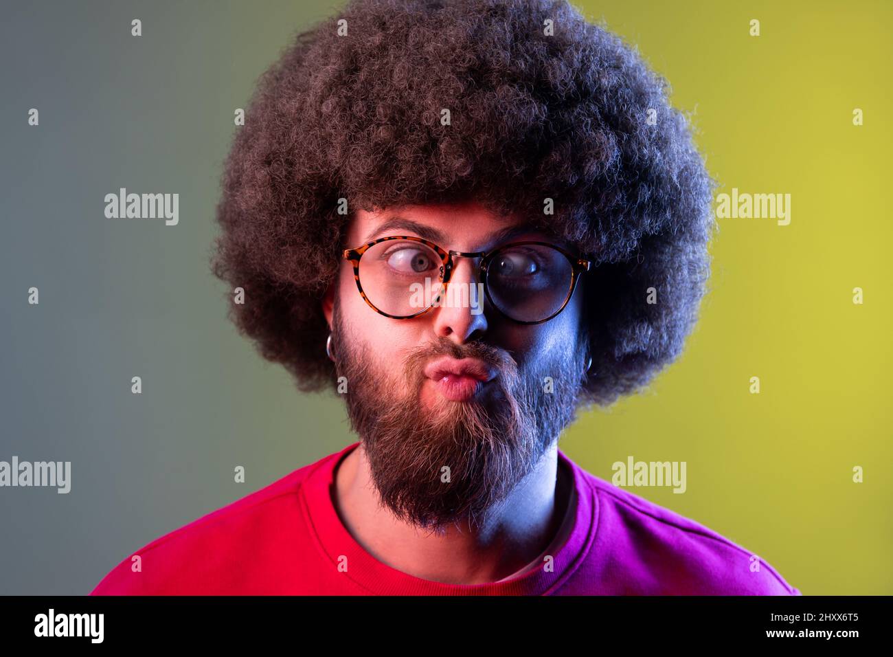 Ritratto di un hipster pazzo con acconciatura afro in piedi con occhi incrociati e guardando con il viso comico divertente, indossando una felpa rossa. Studio interno girato isolato su colorato sfondo di luce al neon Foto Stock