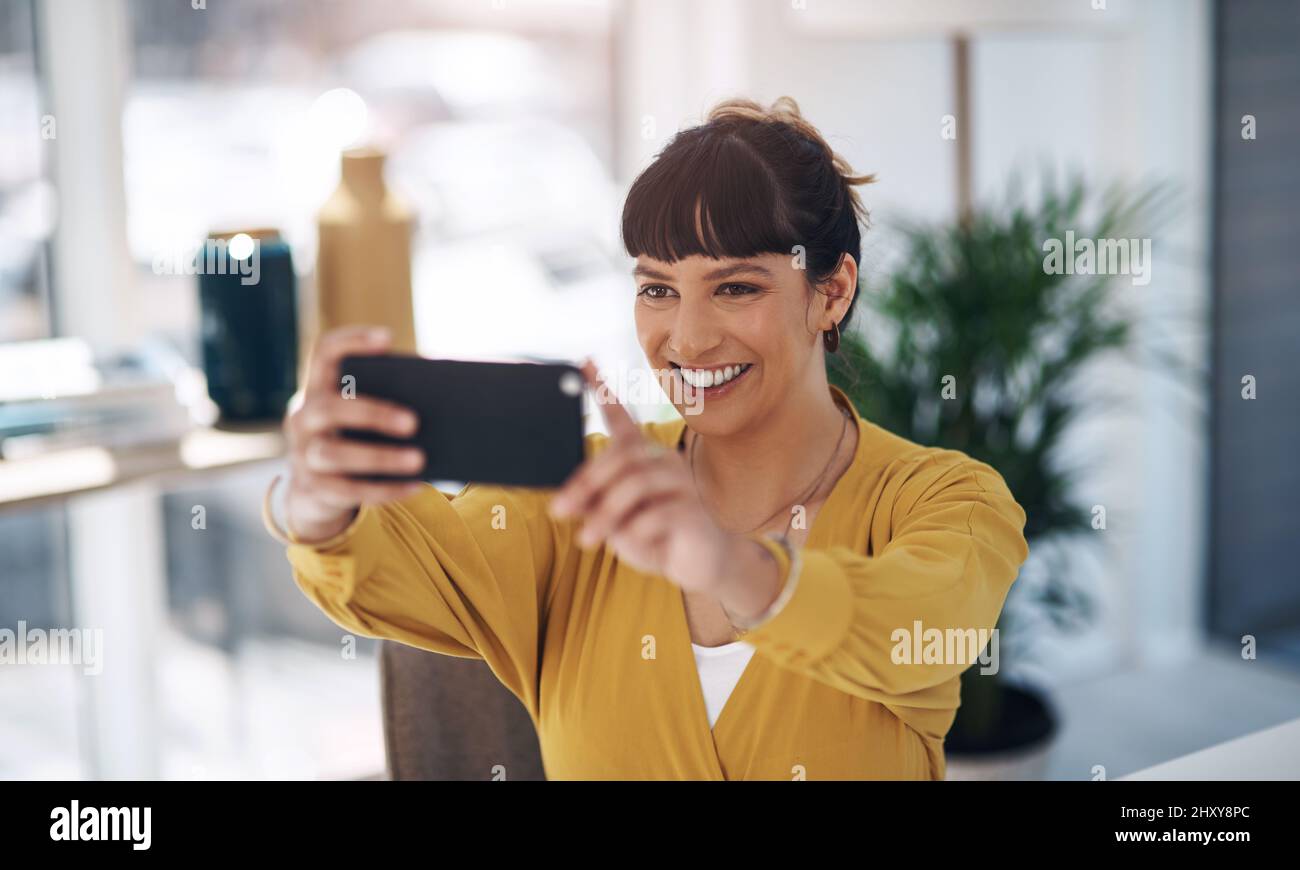 Devo ottenere il mio vestito del giorno. Scatto corto di una giovane donna d'affari attraente seduta da sola nel suo ufficio e usando il suo cellulare per prendere un Foto Stock