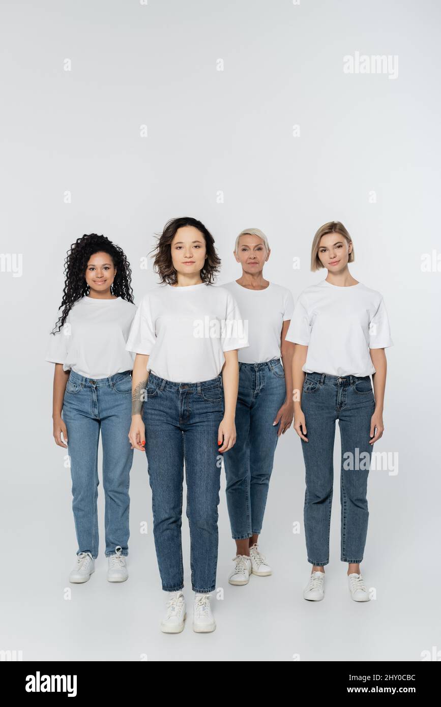 Intera lunghezza di donne multietniche sorridenti in magliette bianche su sfondo grigio, immagine stock Foto Stock