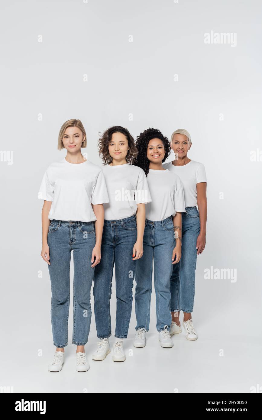 Intera lunghezza di donne interrazziali in jeans e t-shirt su sfondo grigio, femminismo concetto, stock immagine Foto Stock