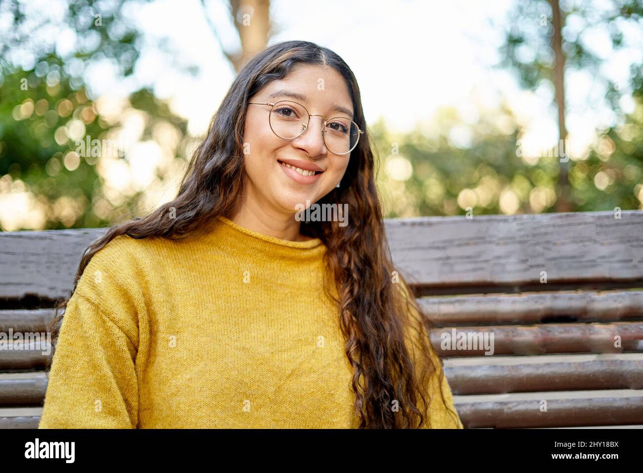 Giovane ragazza allegra con lunghi capelli scuri guardando la macchina fotografica con il sorriso mentre si siede su panca di legno nel parco su sfondo sfocato Foto Stock
