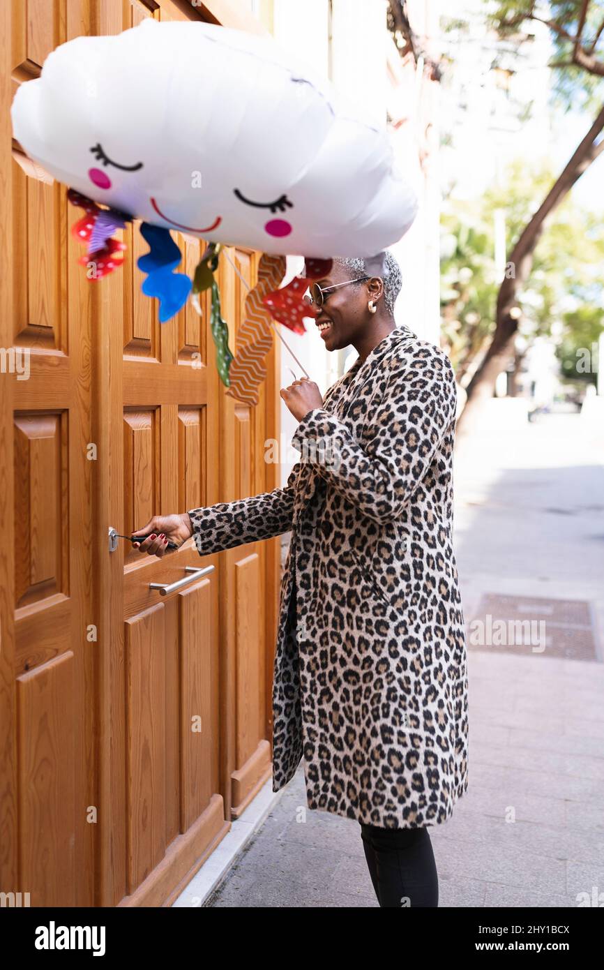 Vista laterale di una femmina afro-americana positiva con palloncino nuvoloso che apre la porta d'ingresso con la chiave mentre si trova sulla strada Foto Stock