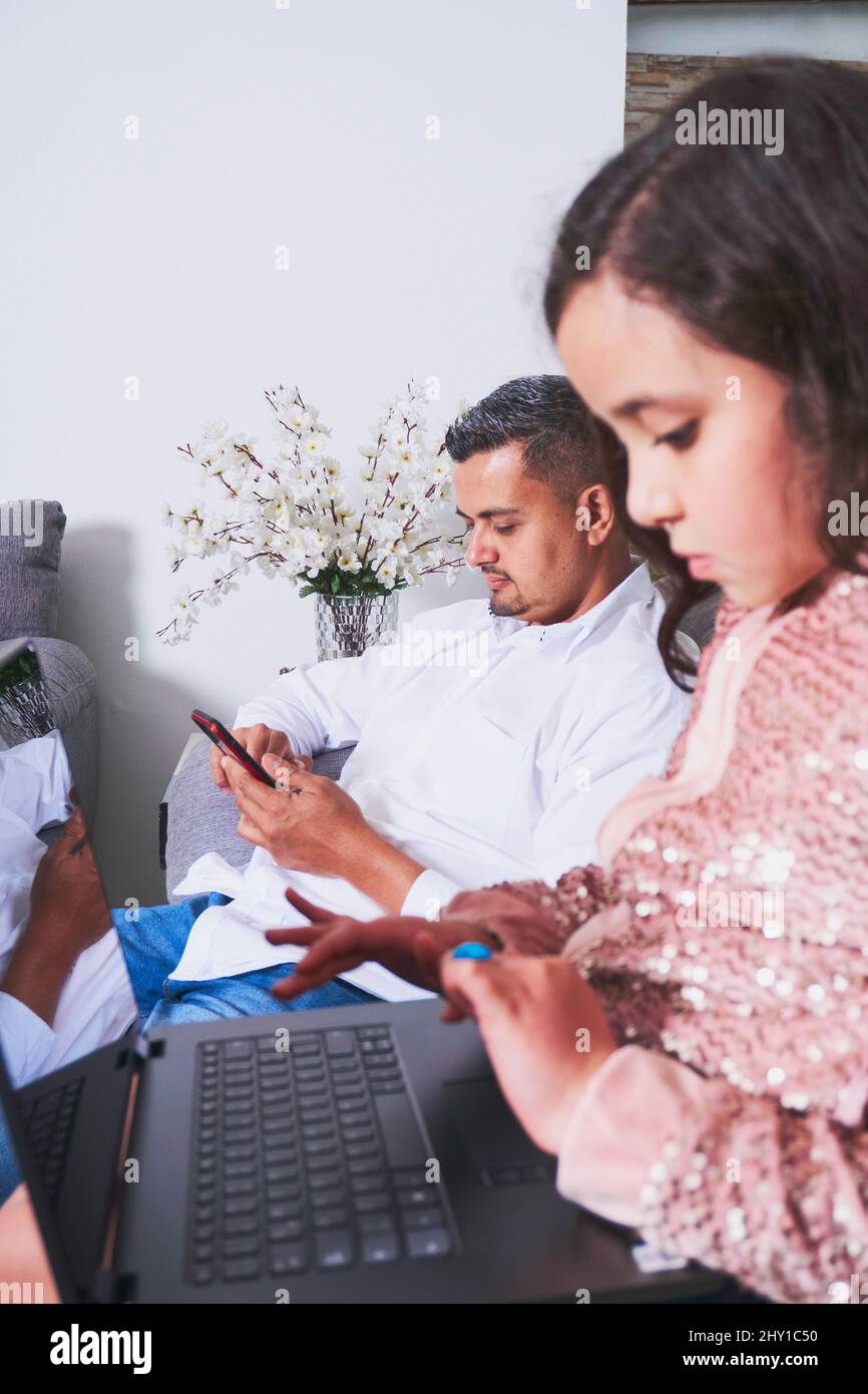 Vista laterale della ragazza ispanica che batte sul netbook mentre si siede sul comodo divano vicino al padre che naviga smartphone in salotto Foto Stock