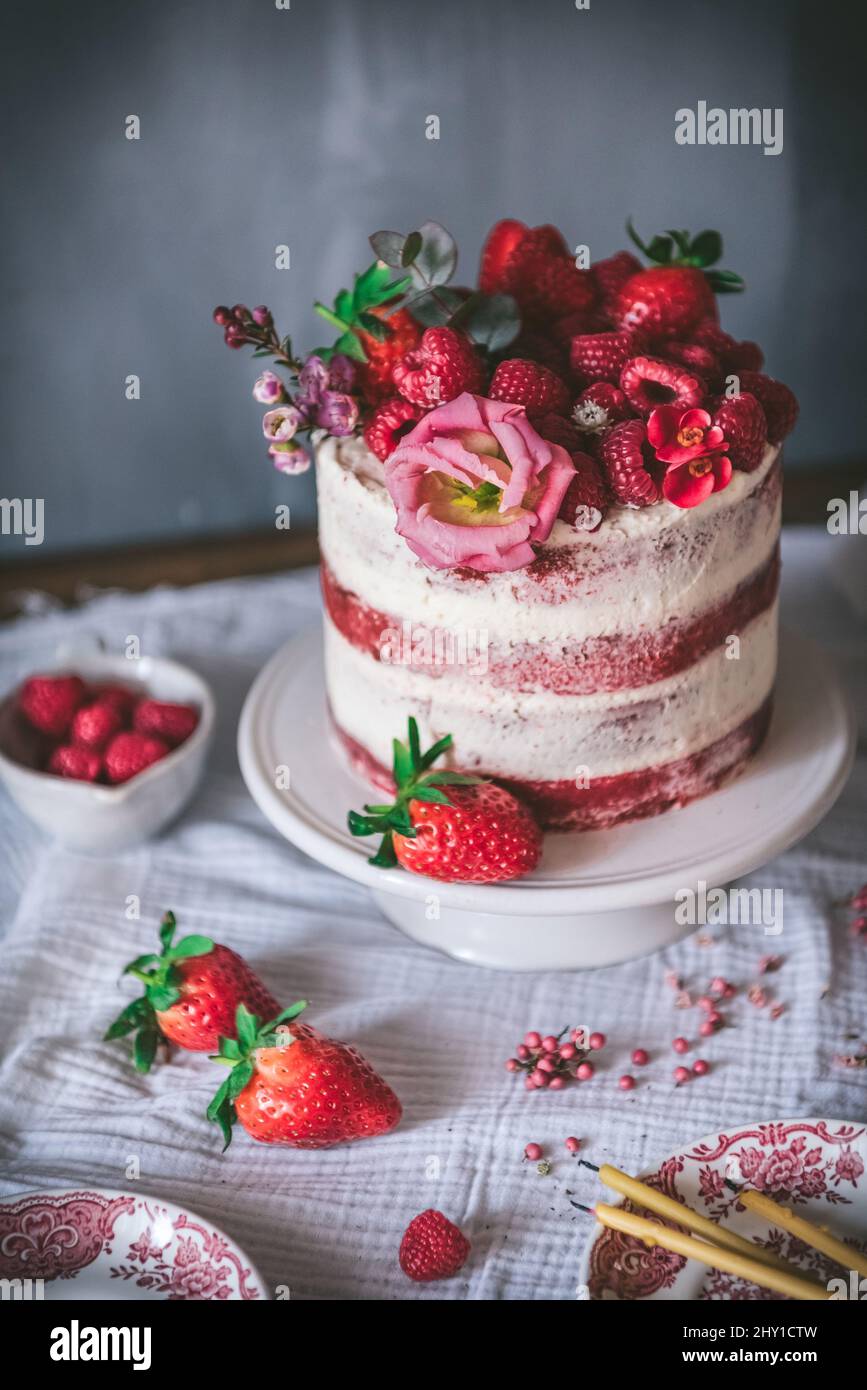 Deliziosa torta di velluto rosso sormontata da fragole fresche e decorata con fiori servita al tavolo con tovaglia bianca in cucina Foto Stock
