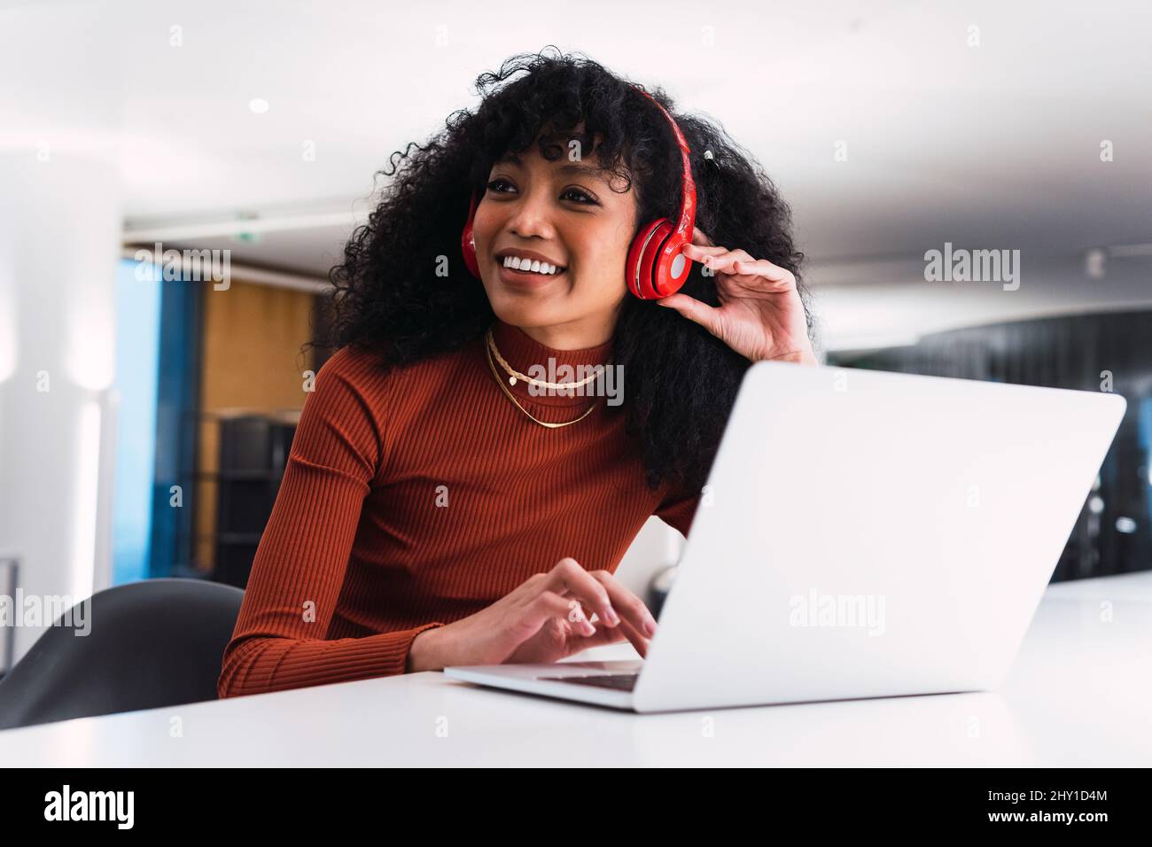 Contenuti afroamericana femminile in cuffie utilizzando il telefono seduto al tavolo con il portatile in un ambiente di lavoro moderno Foto Stock