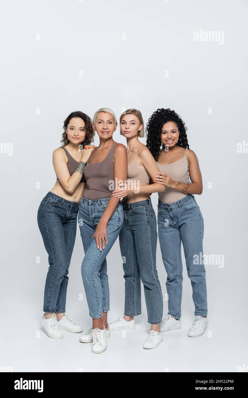 Intera lunghezza di donne multietniche in cima e jeans in posa su sfondo grigio, immagine stock Foto Stock