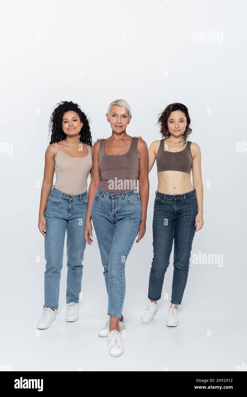 Intera lunghezza di donne multietniche in piedi vicino ad un amico anziano su sfondo grigio, immagine di stock Foto Stock