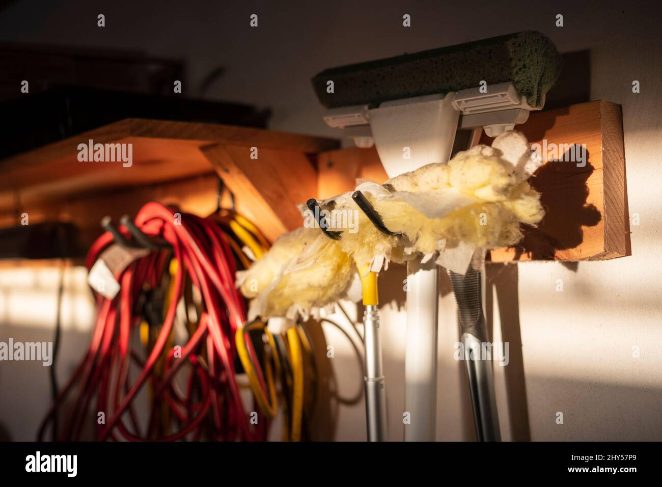 Un piumino di piume è appeso a una parete in un garage accanto alla spugna per mocio e al cavo di servizio come un fascio di luce da un tramonto brilla su di loro. Foto Stock