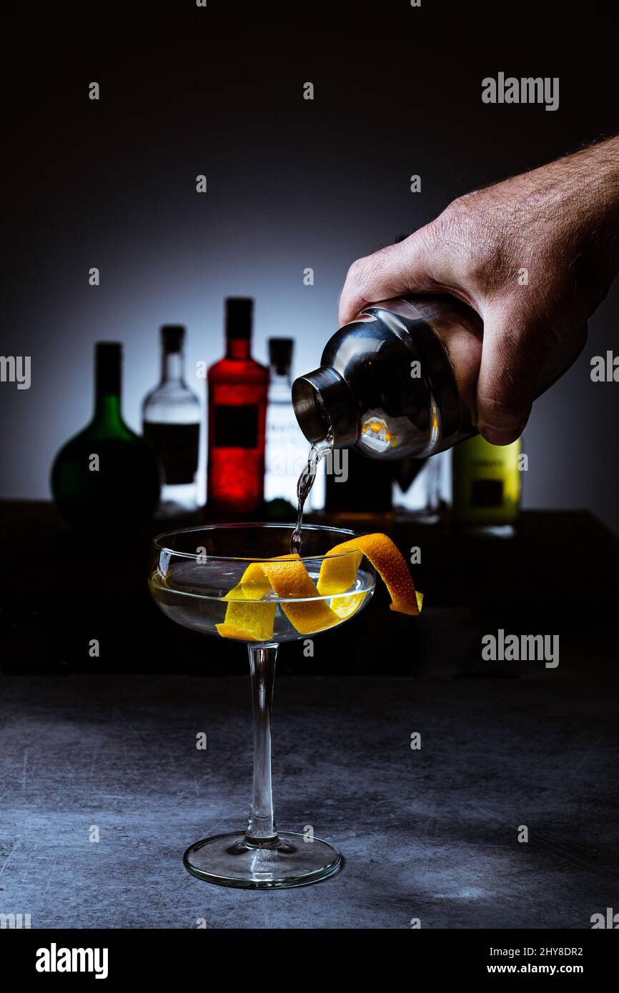 Messa a fuoco poco profonda di una mano che pora Vesper cocktail drink in una tazza di vetro con bottiglie sullo sfondo Foto Stock