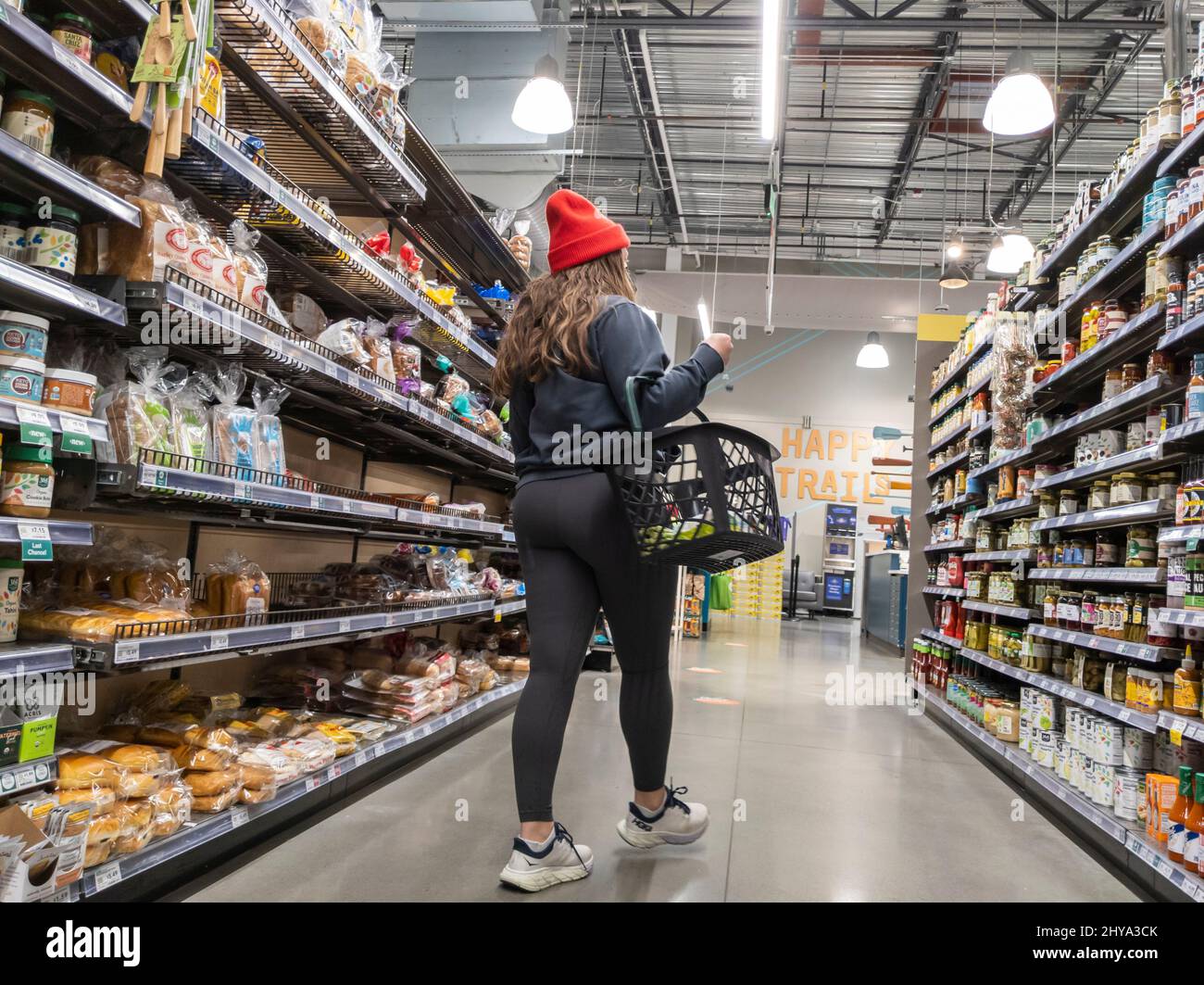 Kirkland, WA USA - circa Febbraio 2022: Donna di Casucasin in un rosso luminoso beanie shopping nel pane e jarred si diffonde corsia all'interno di un intero Foods Mar Foto Stock Kirkland, WA USA - circa Febbraio 2022: Donna di Casucasin in un rosso luminoso beanie shopping nel pane e jarred si diffonde corsia all'interno di un intero Foods Mar Foto Stock