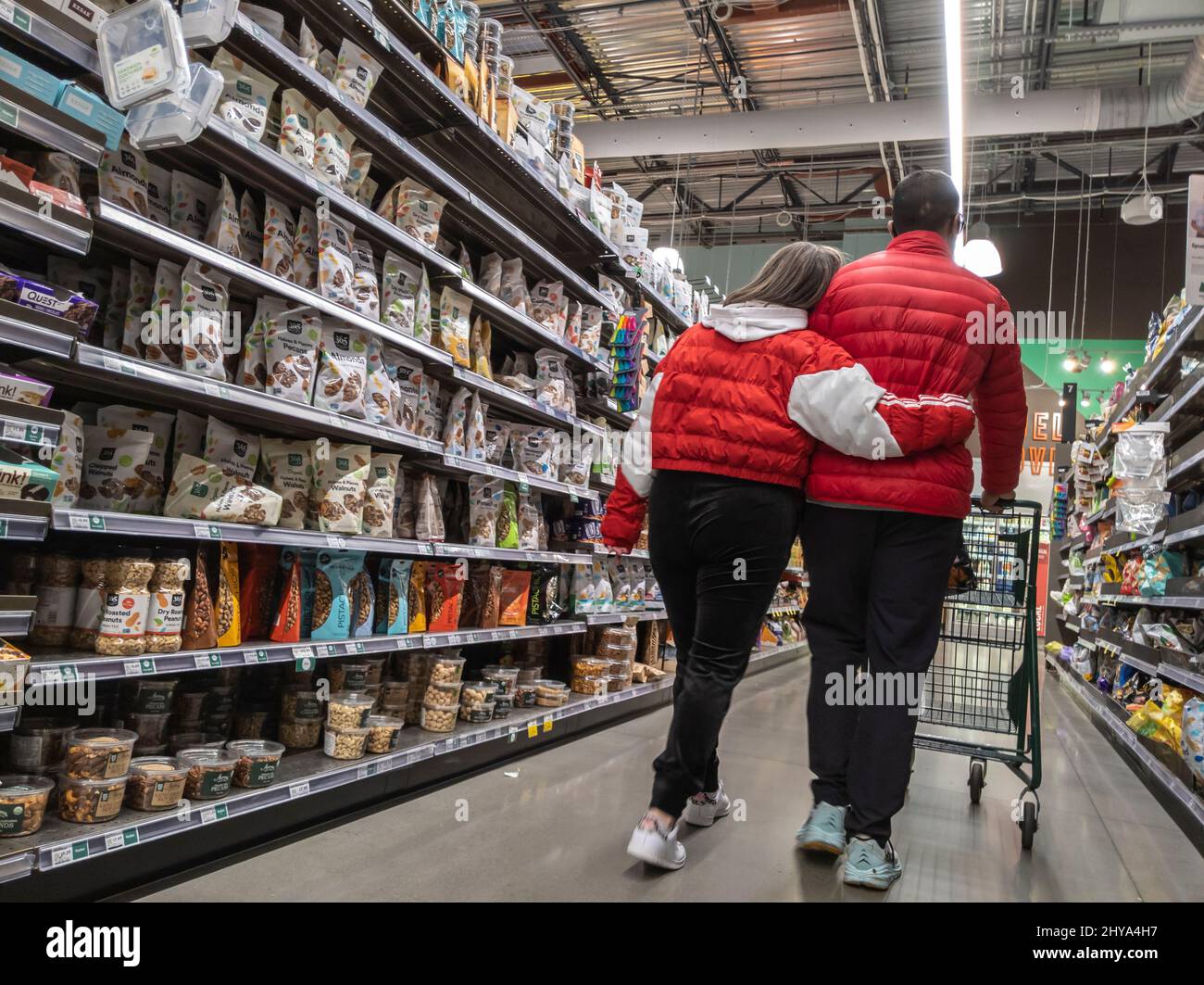 Kirkland, WA USA - circa Febbraio 2022: Vista di una coppia dolce in giacche rosse corrispondenti, che si tengono l'un l'altro come essi curiosare in un intero mercato Foods. Foto Stock Kirkland, WA USA - circa Febbraio 2022: Vista di una coppia dolce in giacche rosse corrispondenti, che si tengono l'un l'altro come essi curiosare in un intero mercato Foods. Foto Stock