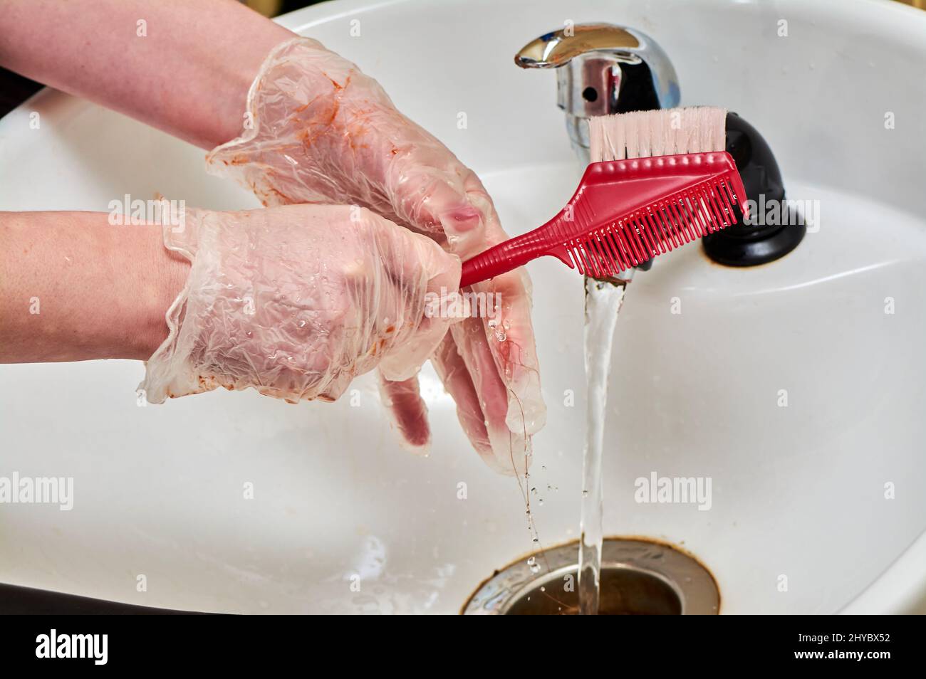 Le mani delle donne nei guanti da colorare dei capelli lavano un pettine sotto un flusso d'acqua in un lavandino bianco Foto Stock