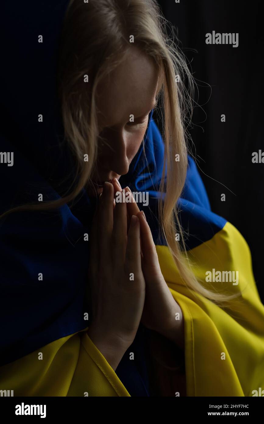 La bionda Ucraina prega con occhi chiusi per l'Ucraina durante la guerra con la bandiera di stato sulla sua testa. La Russia ha attaccato l'Ucraina il 24 febbraio 2022 Foto Stock