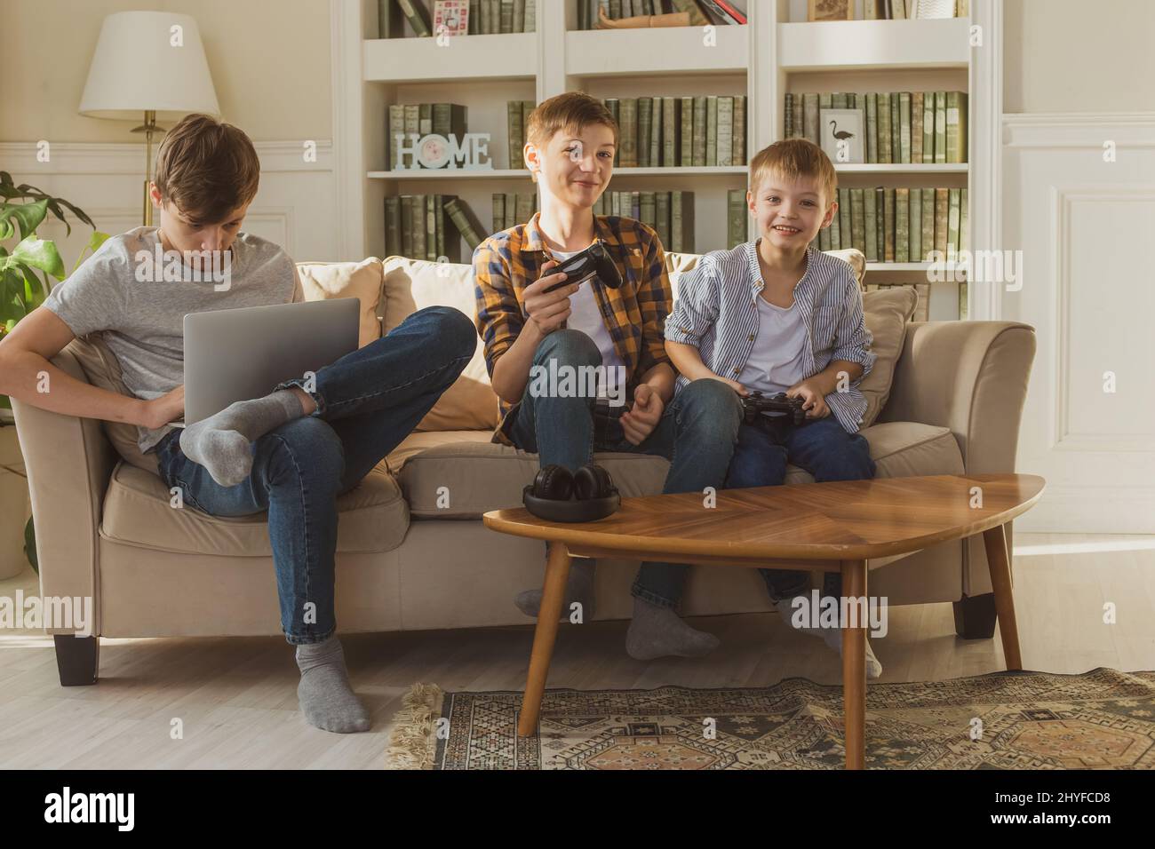 Tre ragazzi, fratelli, si siedono sul divano a casa alla luce del sole e giocano al computer e ai videogiochi. Foto Stock