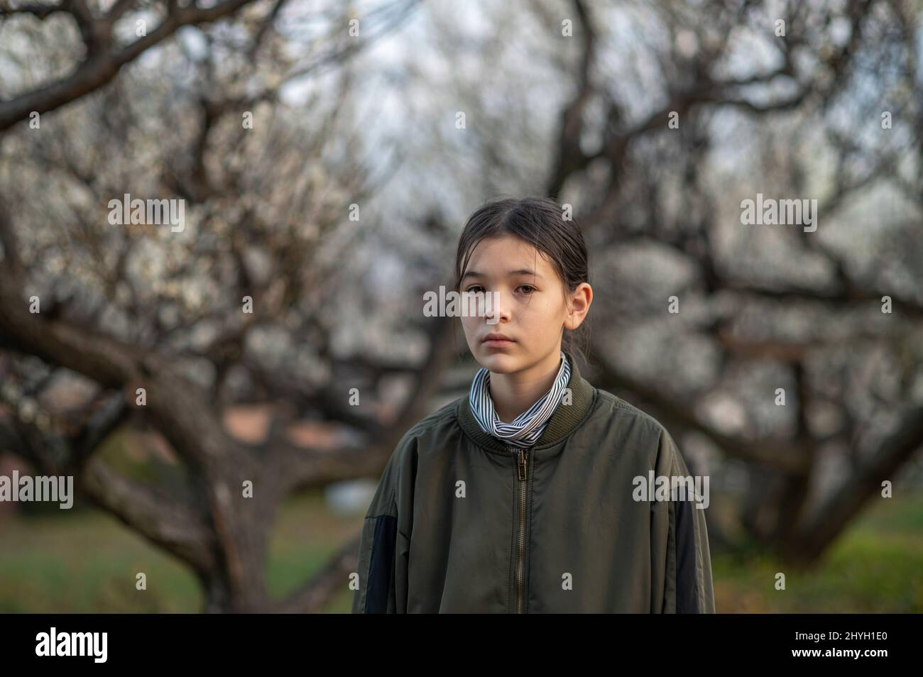 Ritratto di una ragazza preadolescente triste e malata in un parco. Bambino che indossa una giacca verde e un dolcevita. Alberi in fiore sullo sfondo. Primavera. Foto Stock